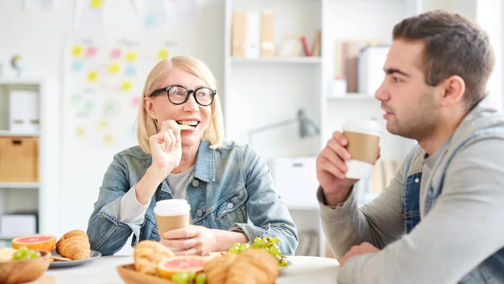 Cheerful coworkers enjoying a coffee break together, eating snacks and chatting in a relaxed office setting.